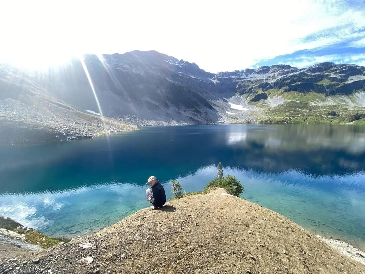 Whistler Olympic Park Hiking in the Callaghan Valley A child sits by a clear, blue mountain lake under a sunny sky.