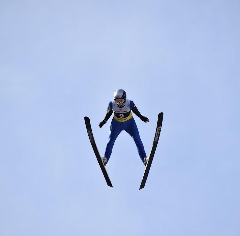 Dyllon Crawford Ski jumper in mid-air against a clear blue sky.