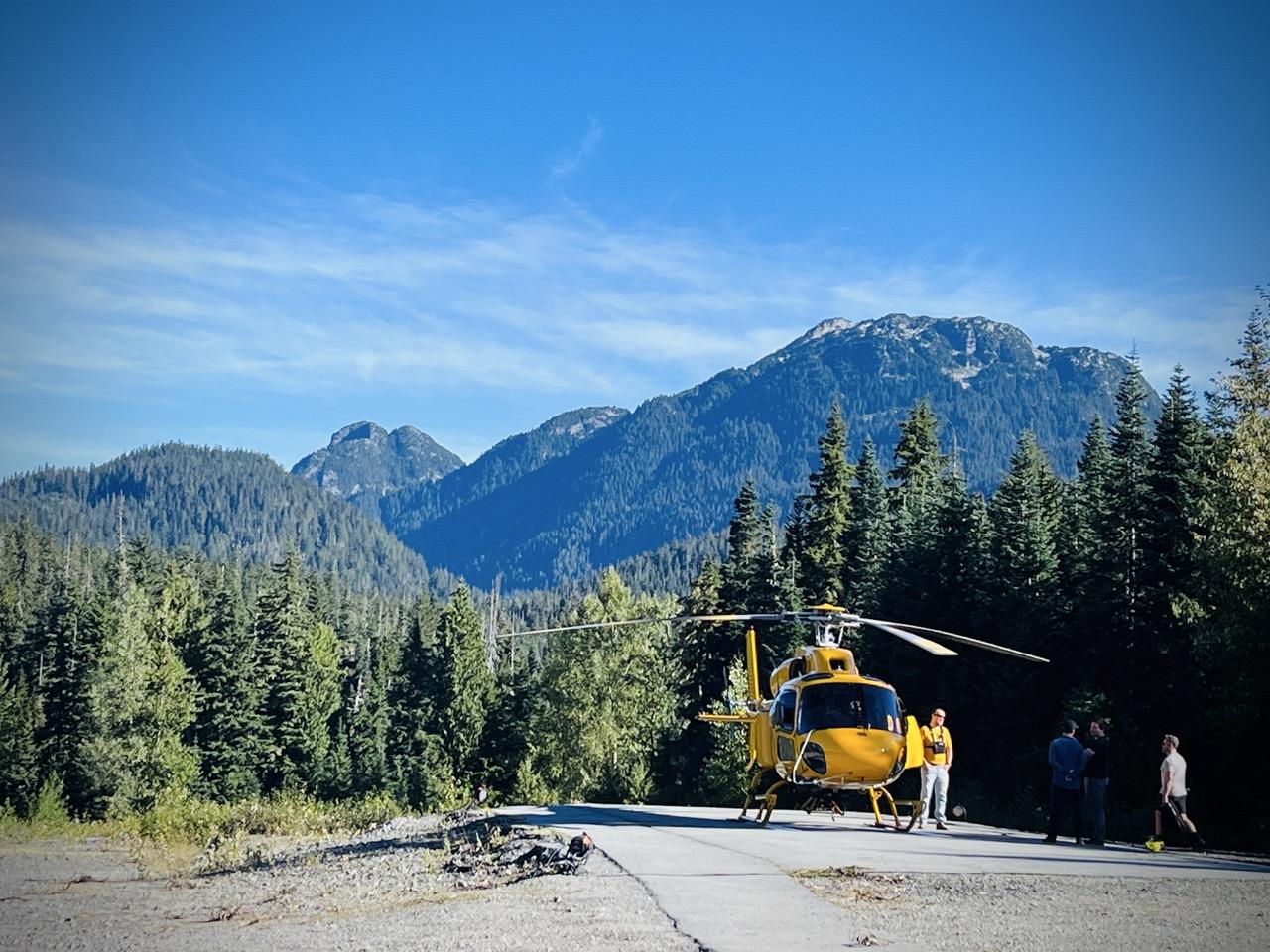 Yellow helicopter on a mountain road, surrounded by pine trees and blue sky.