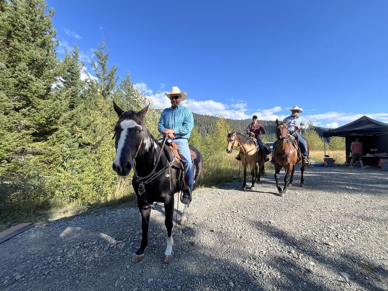 Three people on horses on a gravel path, surrounded by trees on a sunny day.