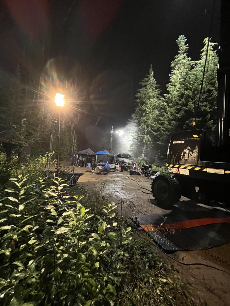 Nighttime forest scene with tents, bright lights, and construction equipment.