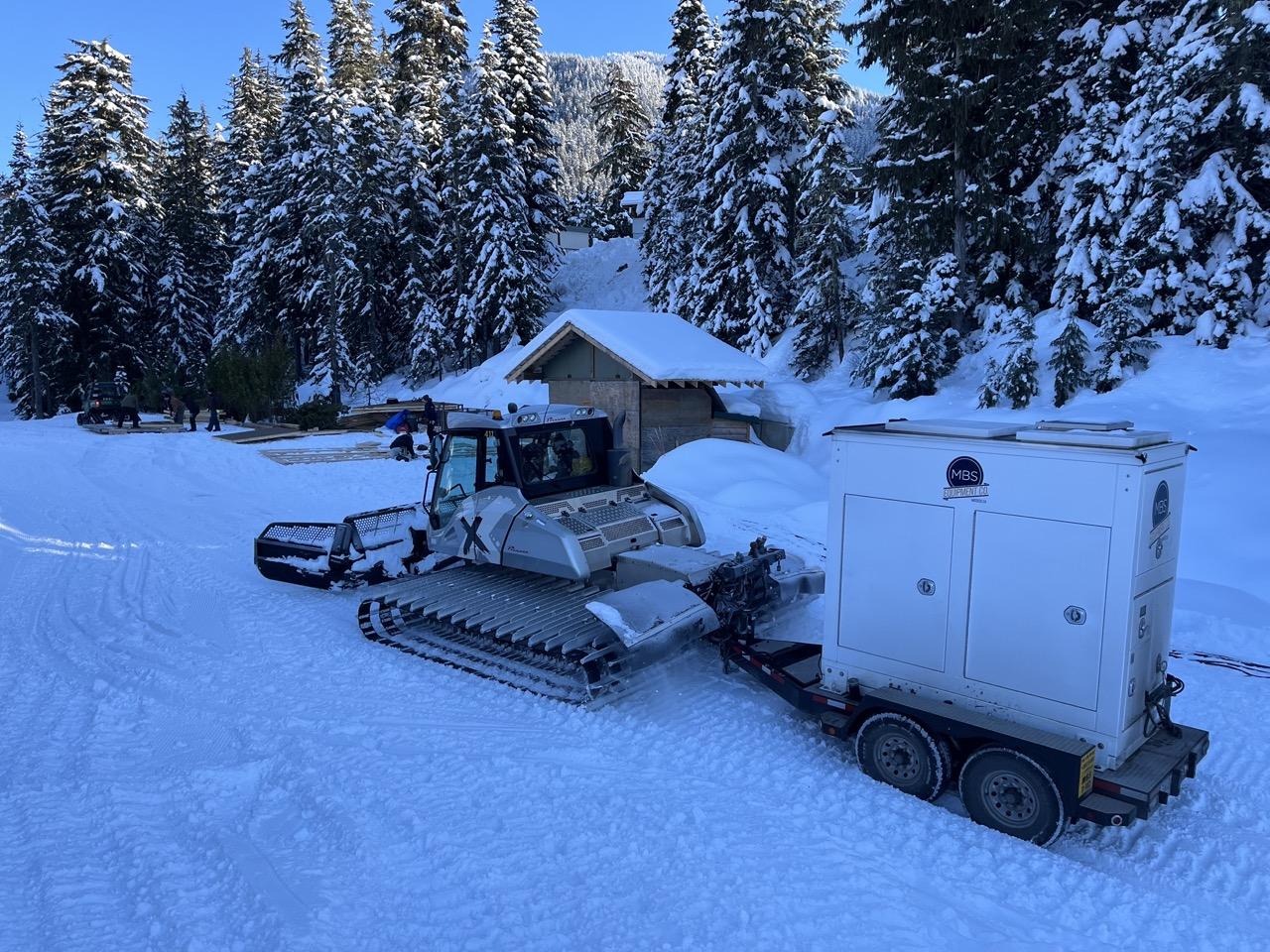 Snow groomer towing a trailer in a snowy forest setting.