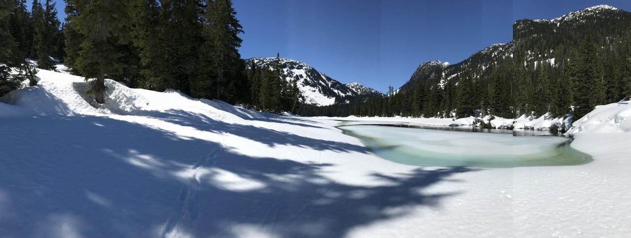 Snowy landscape with frozen lake, surrounded by pine trees under a clear blue sky.