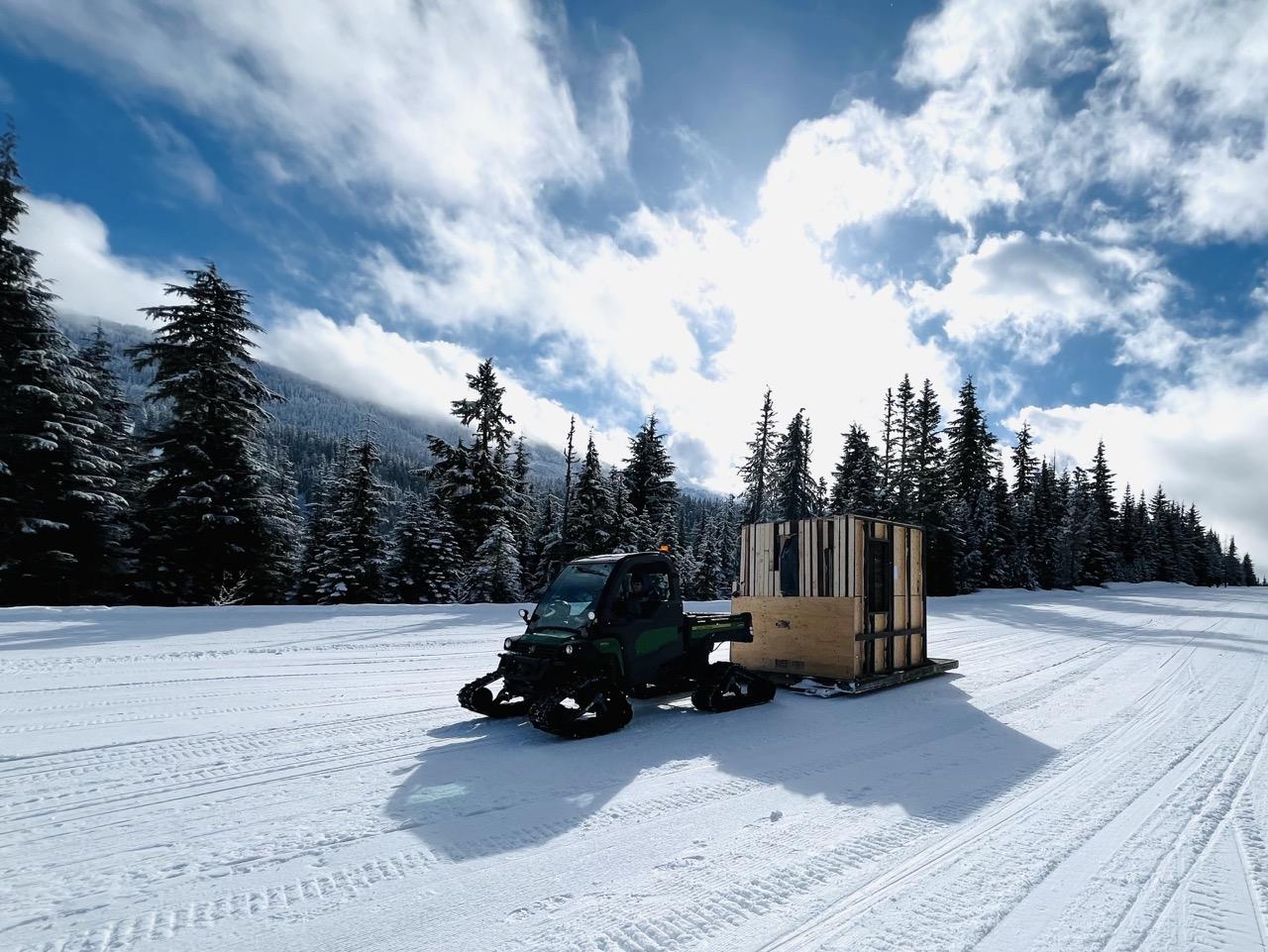 Snowmobile towing a wooden structure on a snowy field under a clear blue sky.