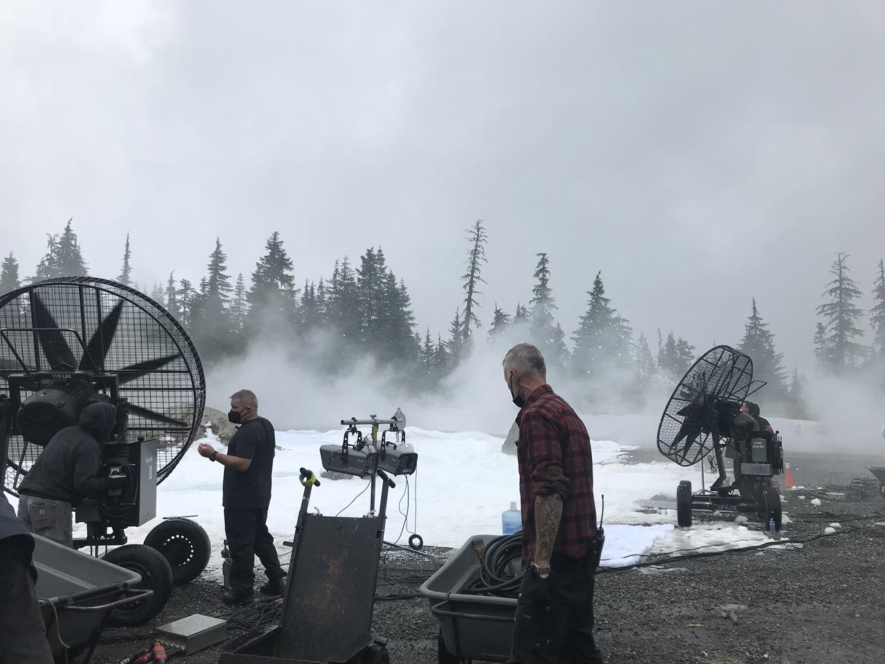 Film crew with large fans in a foggy forest setting.