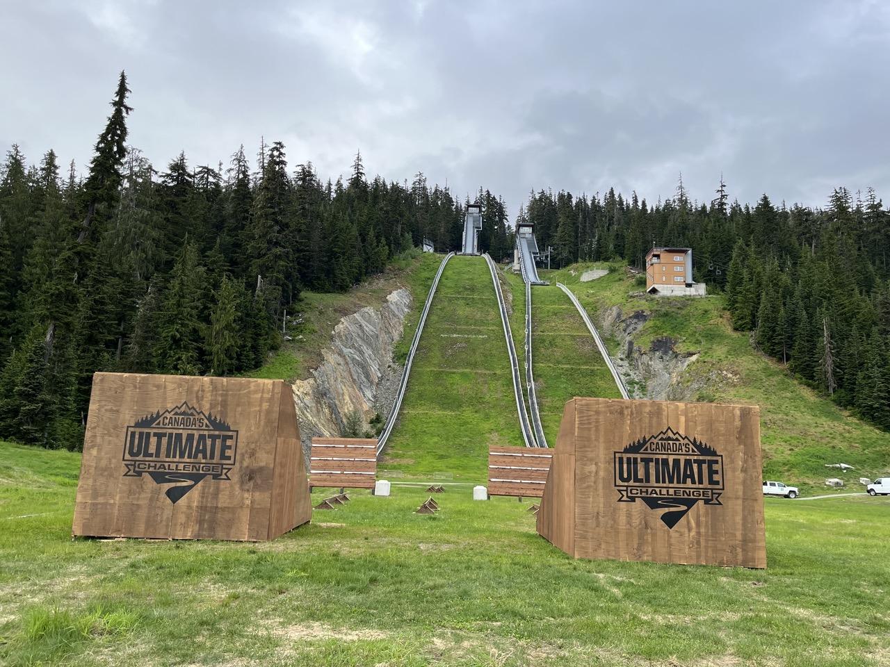 Grassy ski jump hill with wooden signage, surrounded by tall trees.