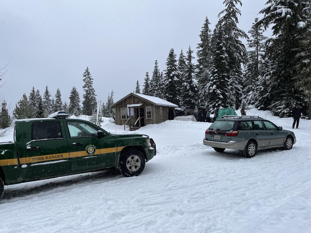 Green park ranger truck and car in snowy forest, cabin in the background.