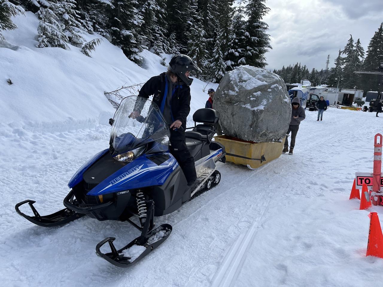 Snowmobile towing a large boulder on a snowy road with trees in the background.