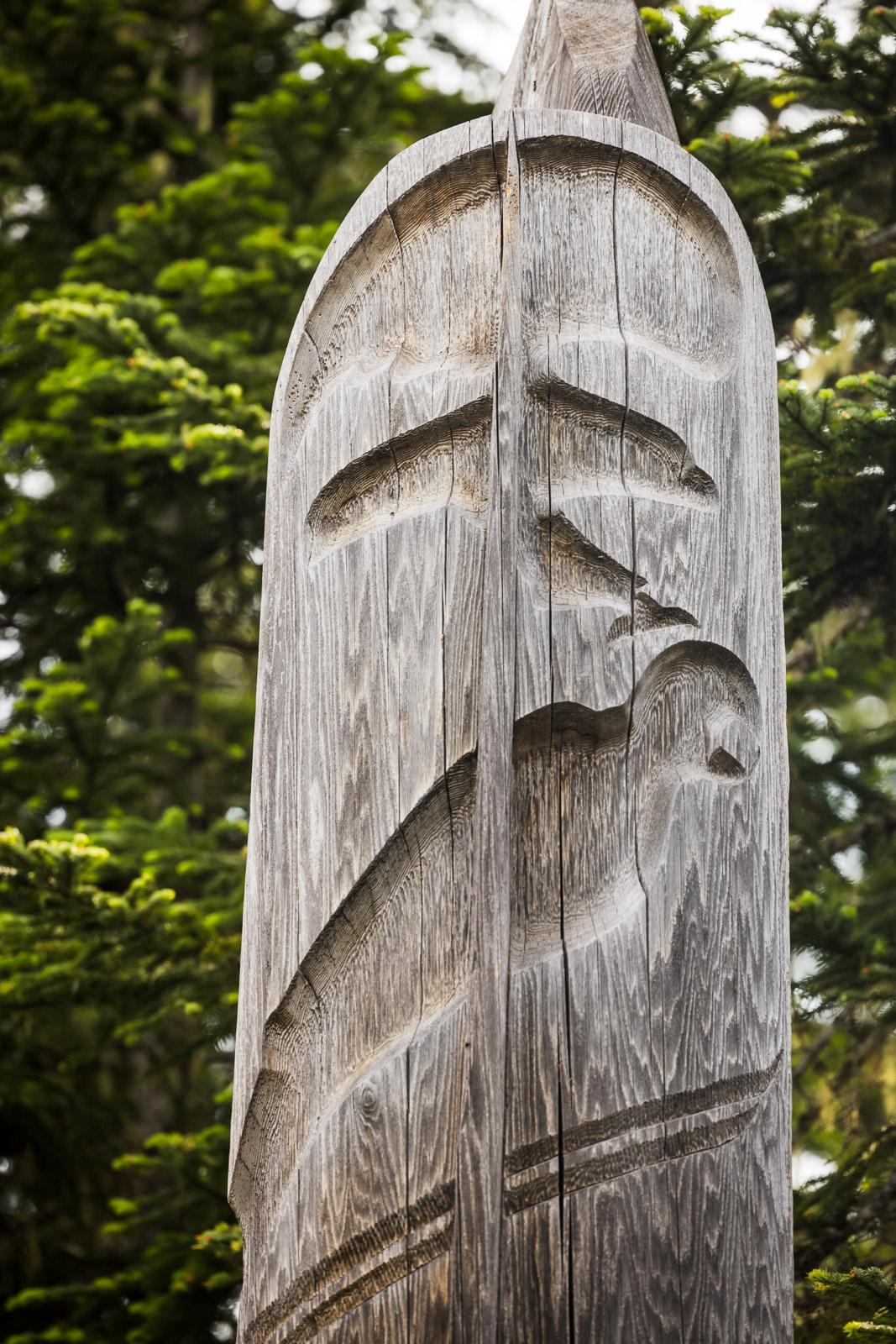 Carved wooden totem pole with abstract face against green trees.