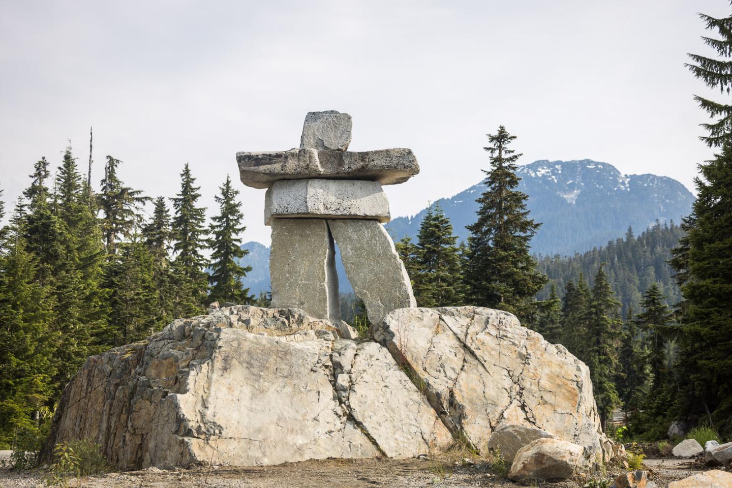 Stone inukshuk on a rocky hill, with evergreen trees and mountains in the background.