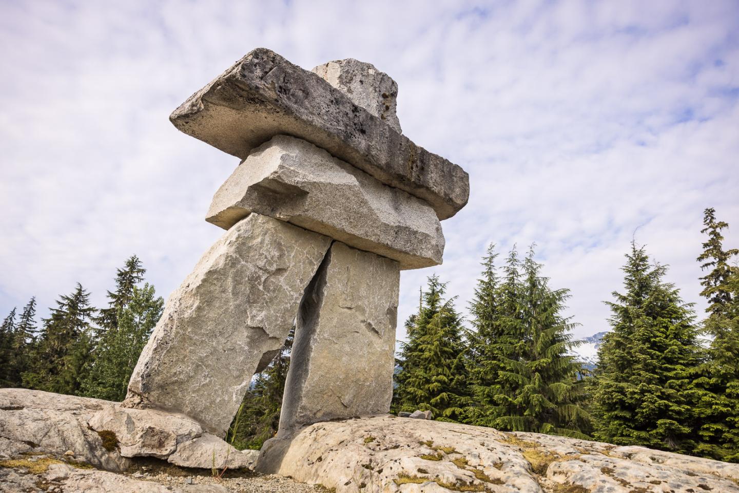 Stone inukshuk sculpture against a forested backdrop and cloudy sky.