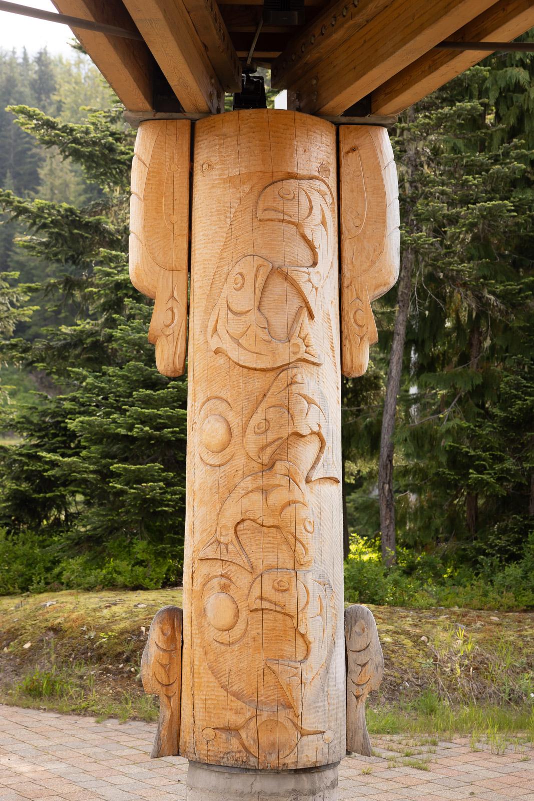 Carved wooden totem pole under a wooden structure, surrounded by evergreen trees.