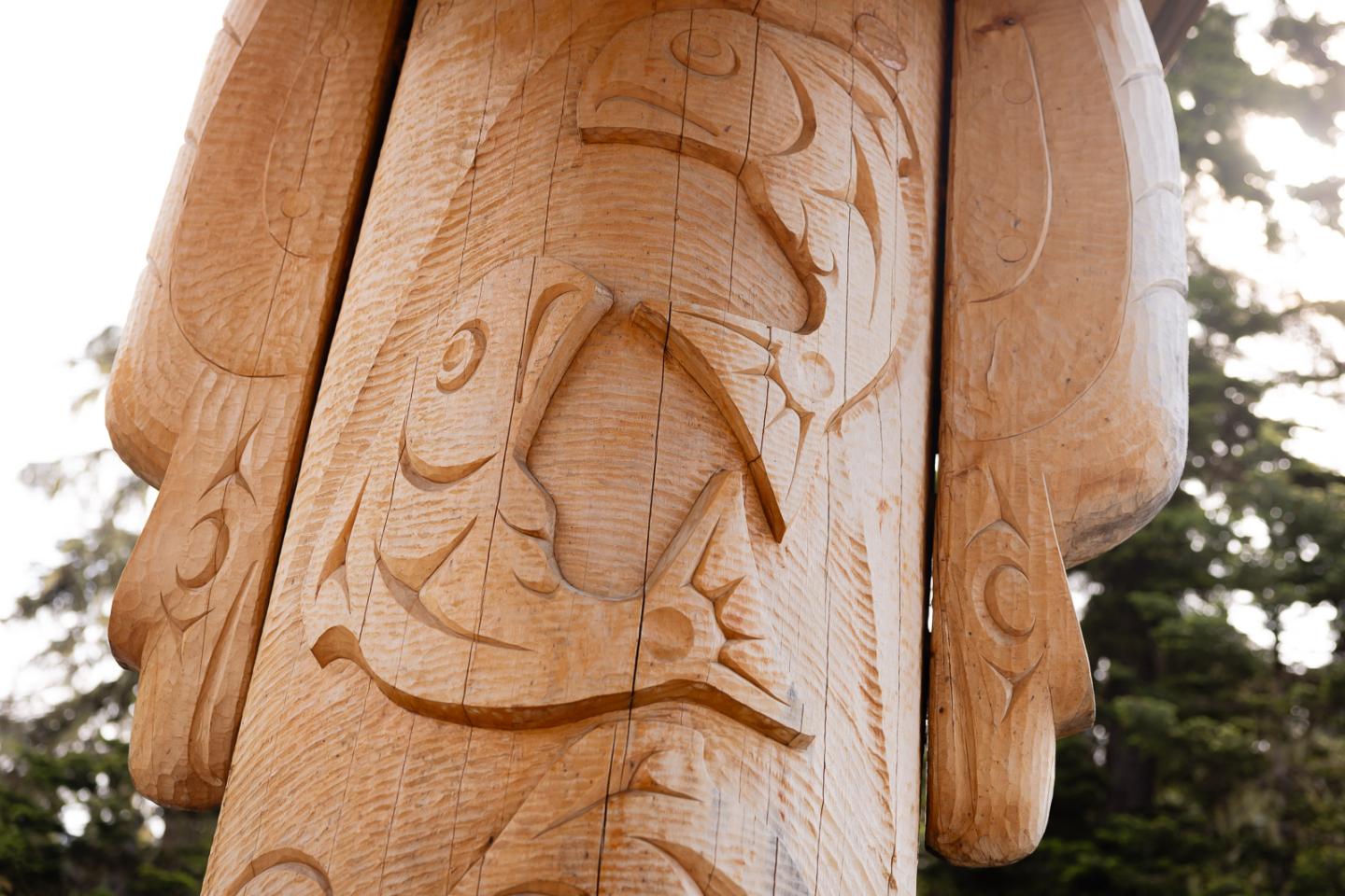Carved totem pole with fish designs, set against trees.