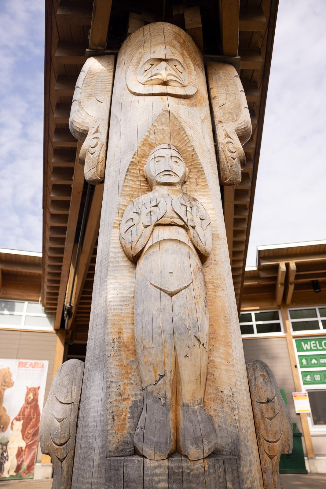 Wooden totem pole with carvings of human figures under a building's eaves.