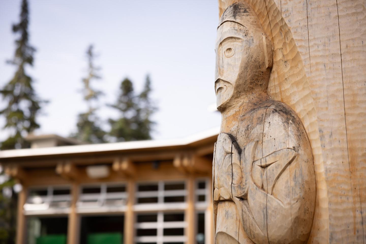 Carved wooden totem pole with building and trees in background.