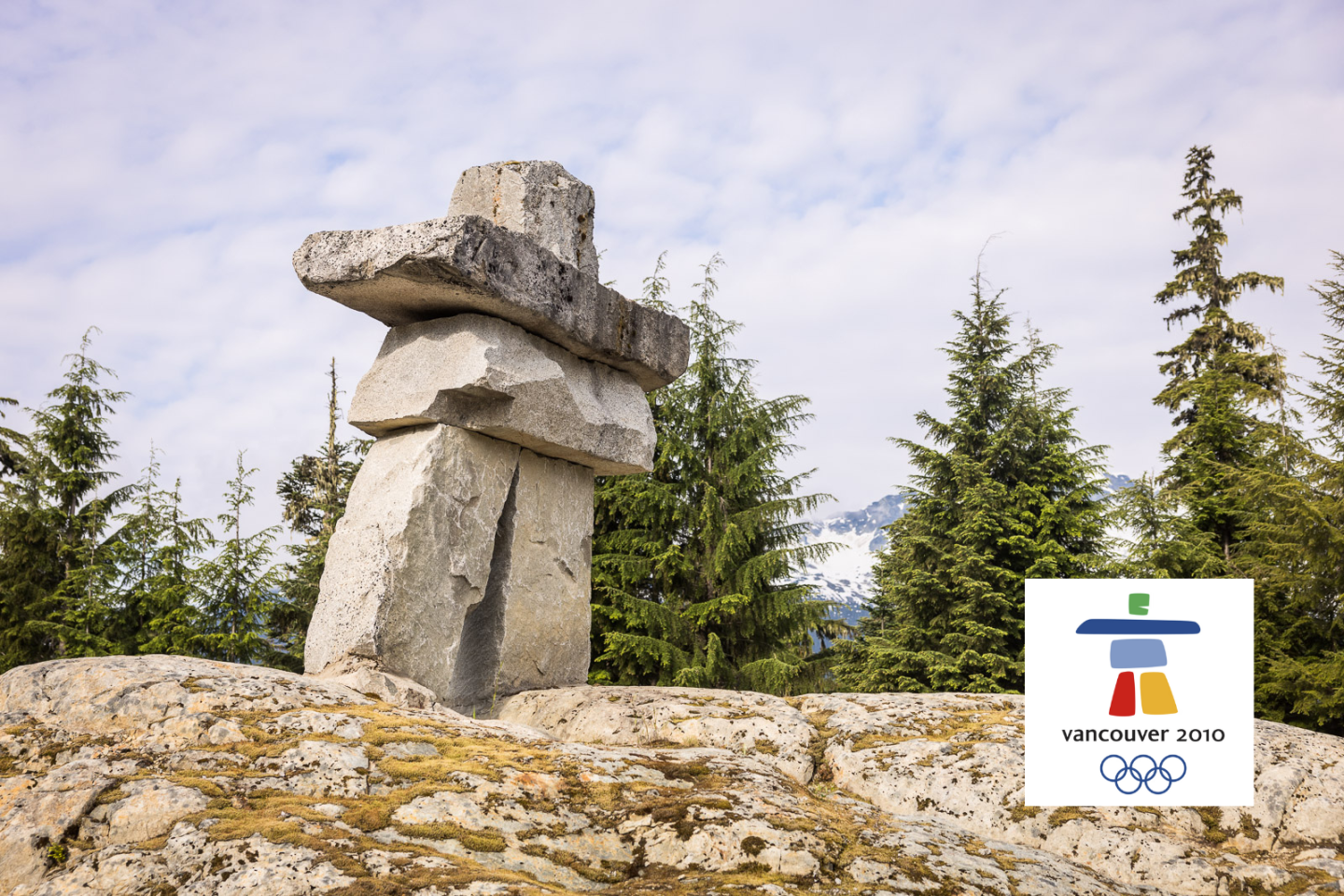 Stone inukshuk with evergreen trees, overcast sky, and Vancouver 2010 logo.
