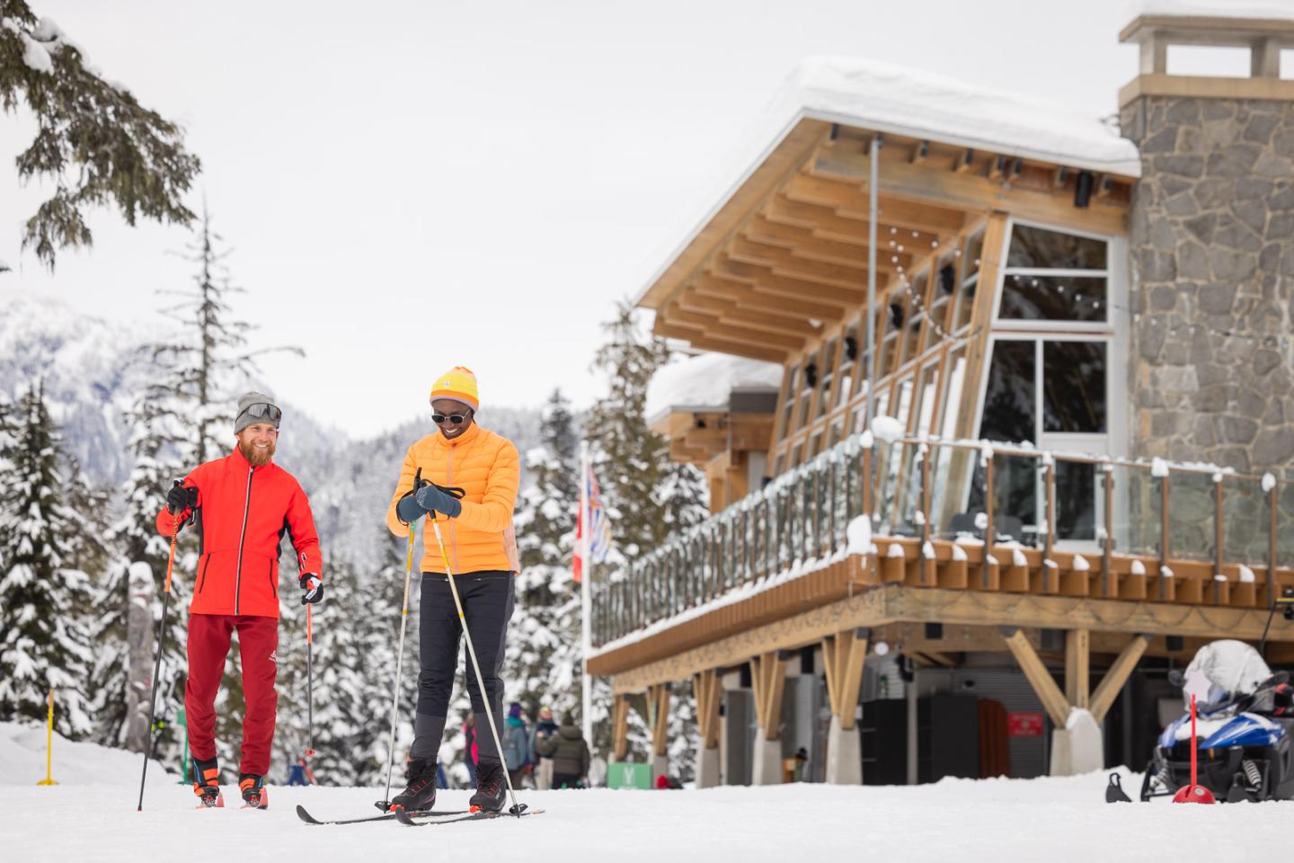 Two people cross-country skiing near a snowy lodge. Whistler Olympic Park