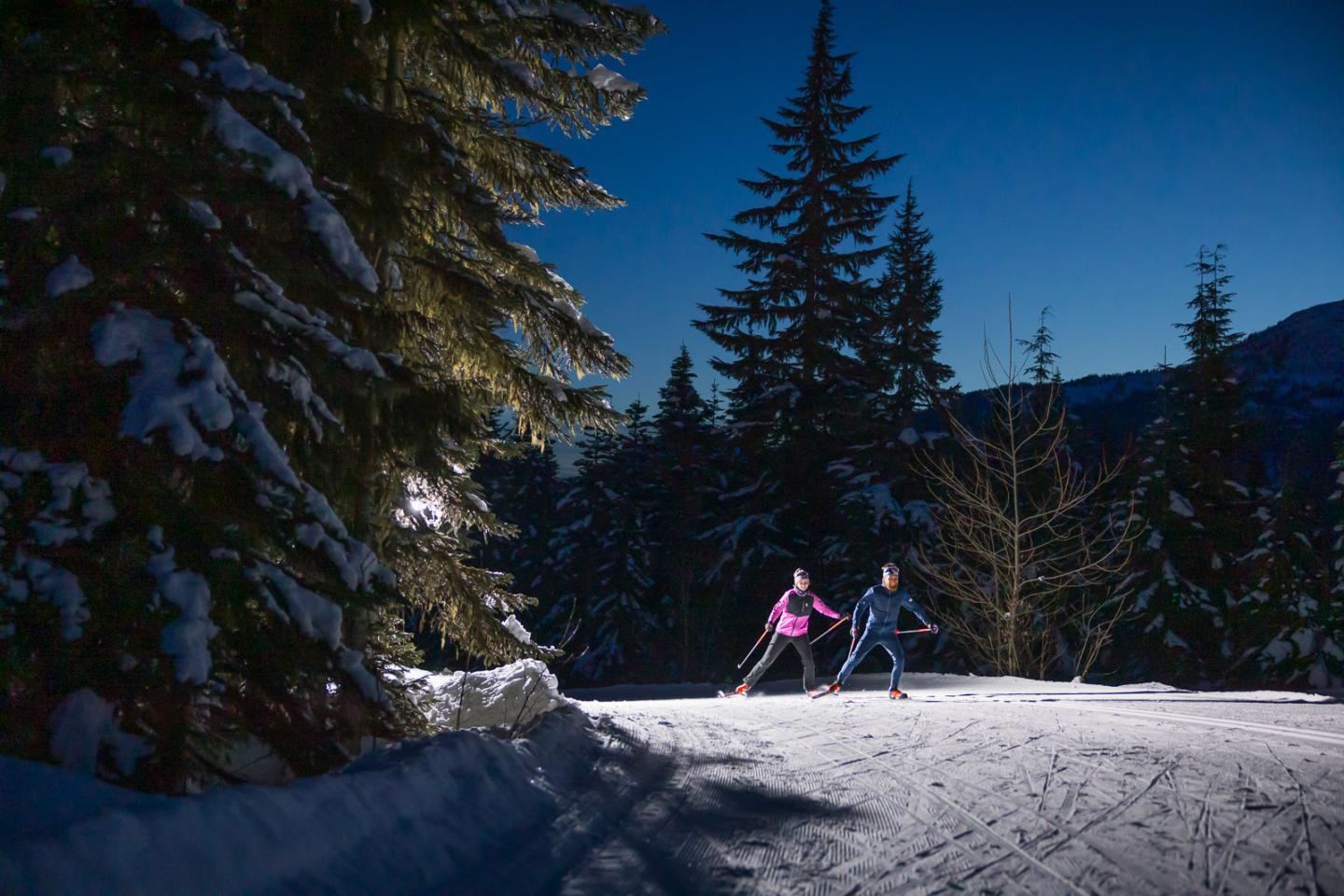 Two people cross-country skiing at night in a snowy forest at Whistler Olympic Park
