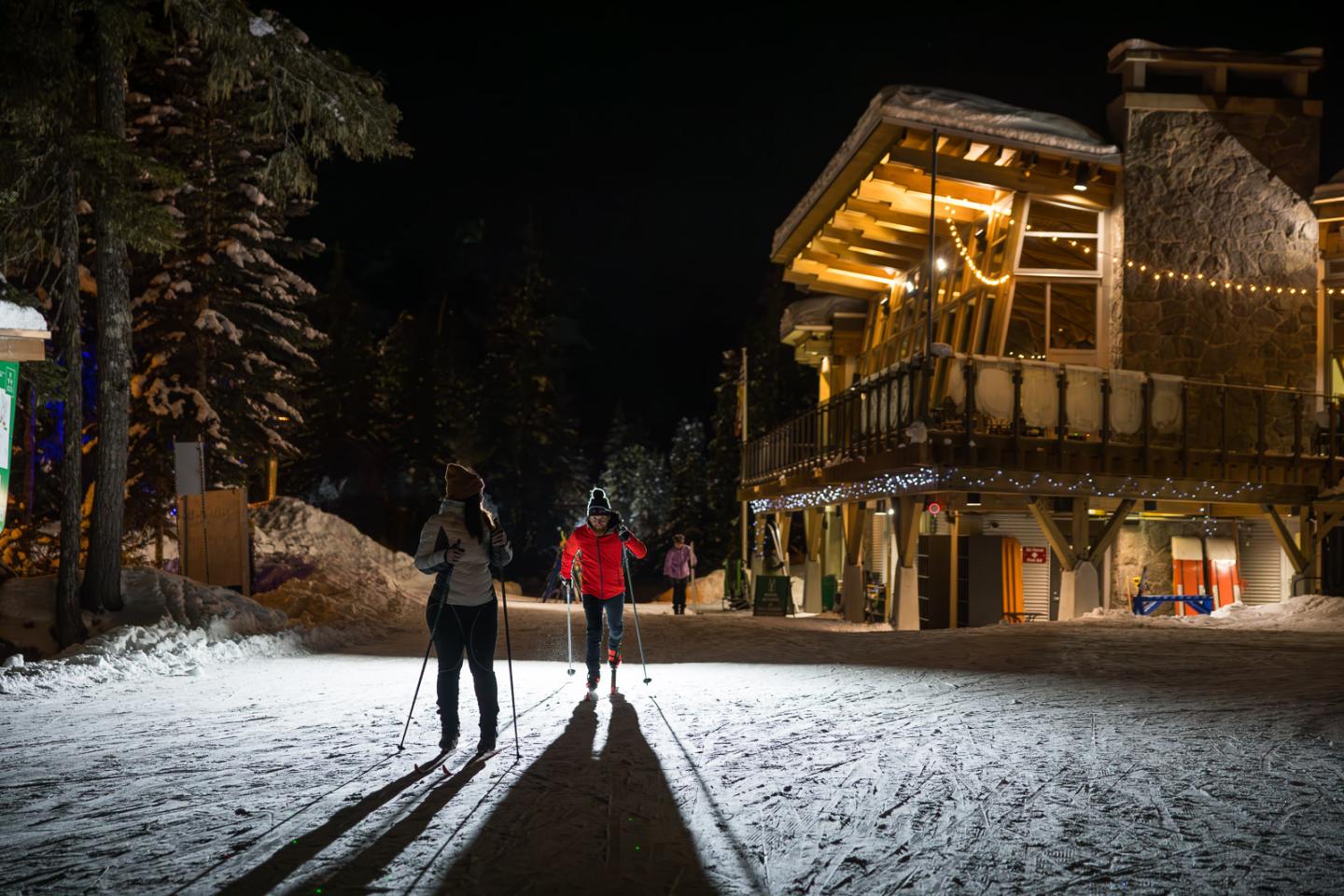 Skiers at night near a lit wooden lodge in a snowy forest at Whistler Olympic Park