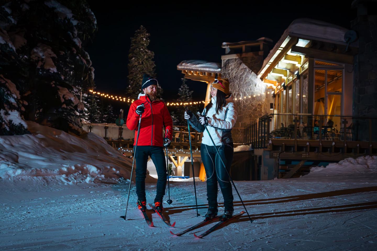 Two people skiing at night near a lit cabin in a snowy setting.