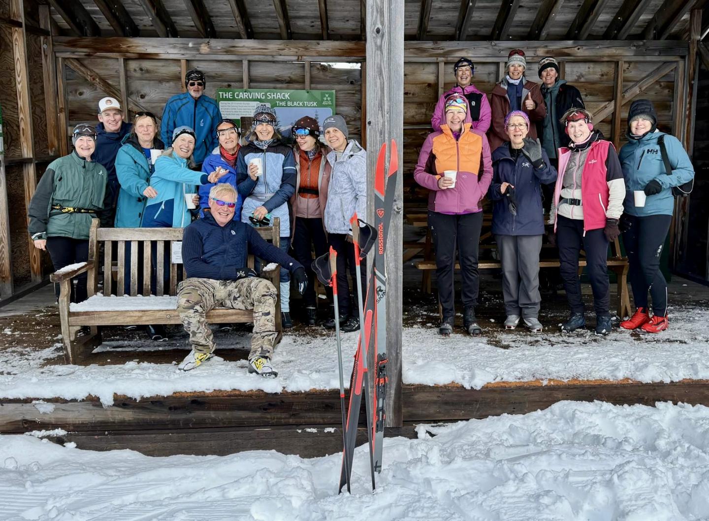 Group of people in winter clothing, smiling in a snowy wooden shelter.