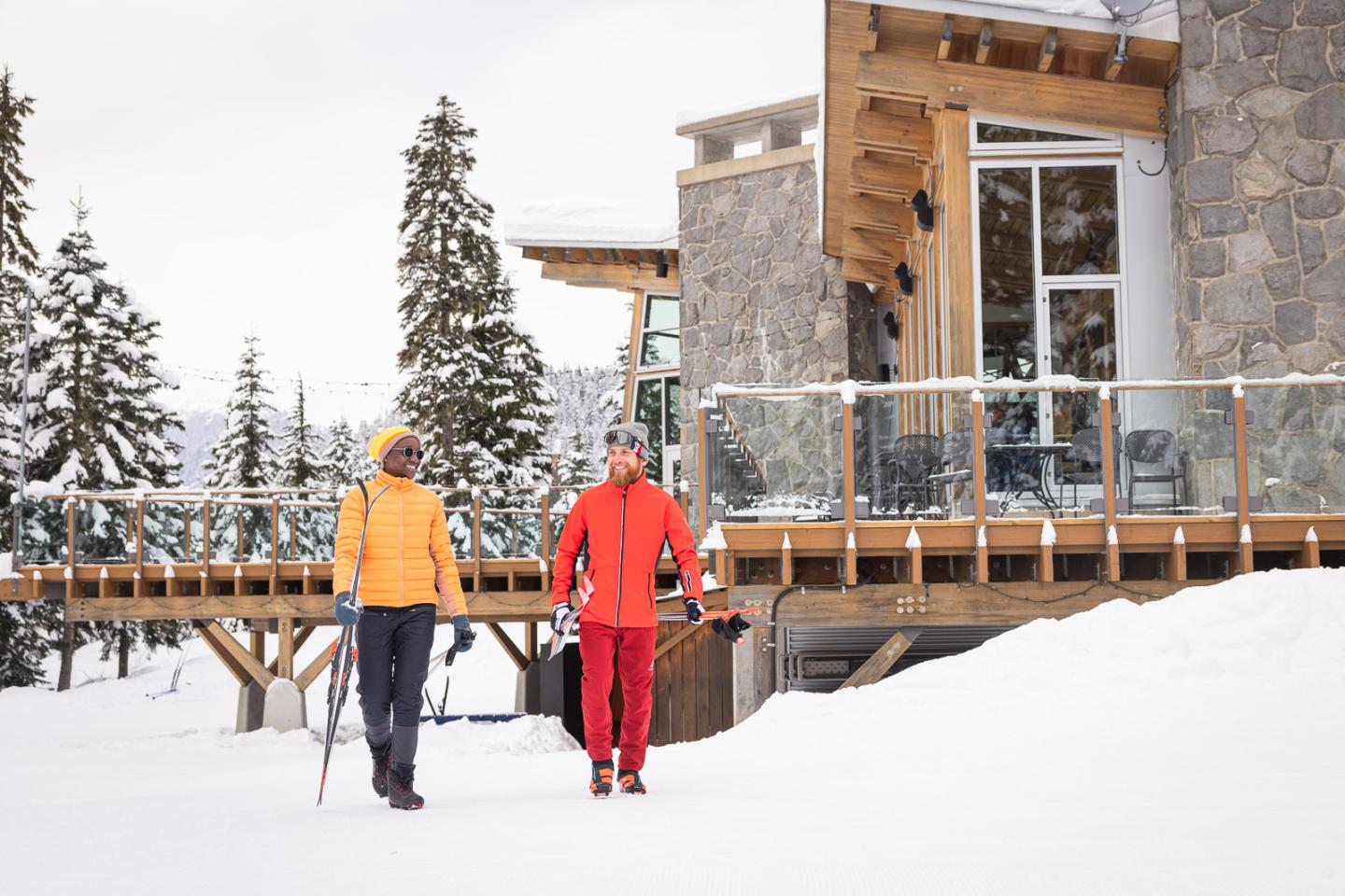 Two people in bright ski gear walk near a snowy cabin.