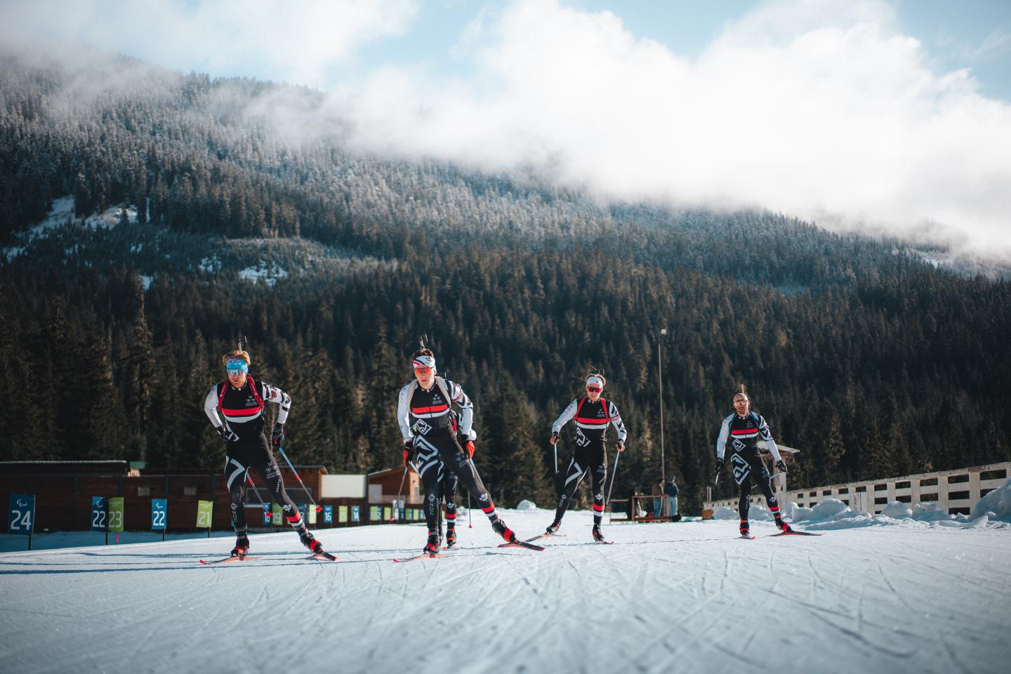 Four Biathlon Athlete skiing Whistler Nordic Development Centre in motion on a snowy trail, with forested mountains in the background.