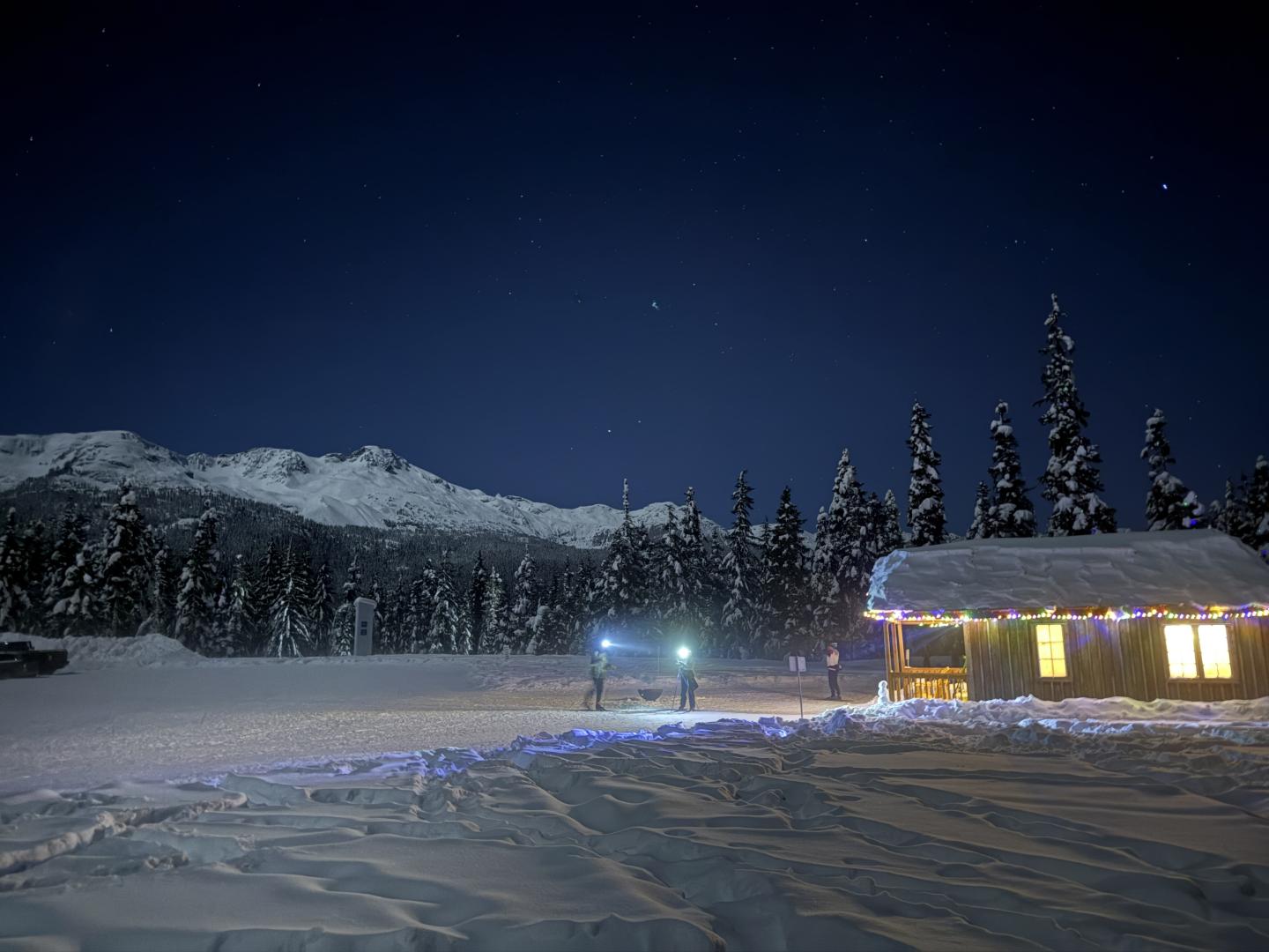 Snowy cabin with colorful lights at night, mountains in the background.