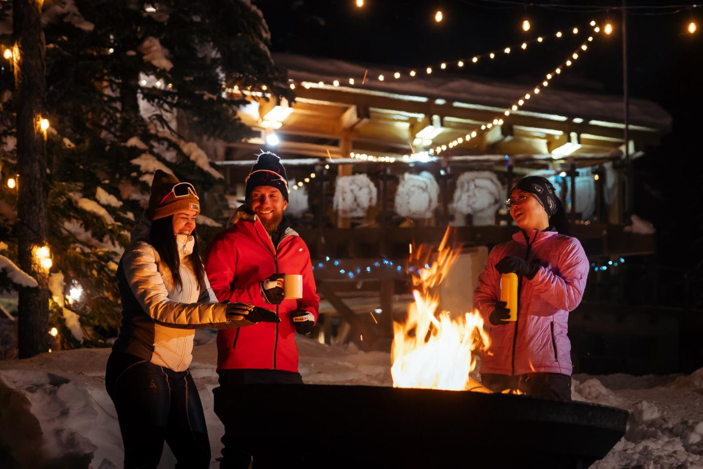 Three people in winter gear enjoying a campfire at night with a chalet in the background.