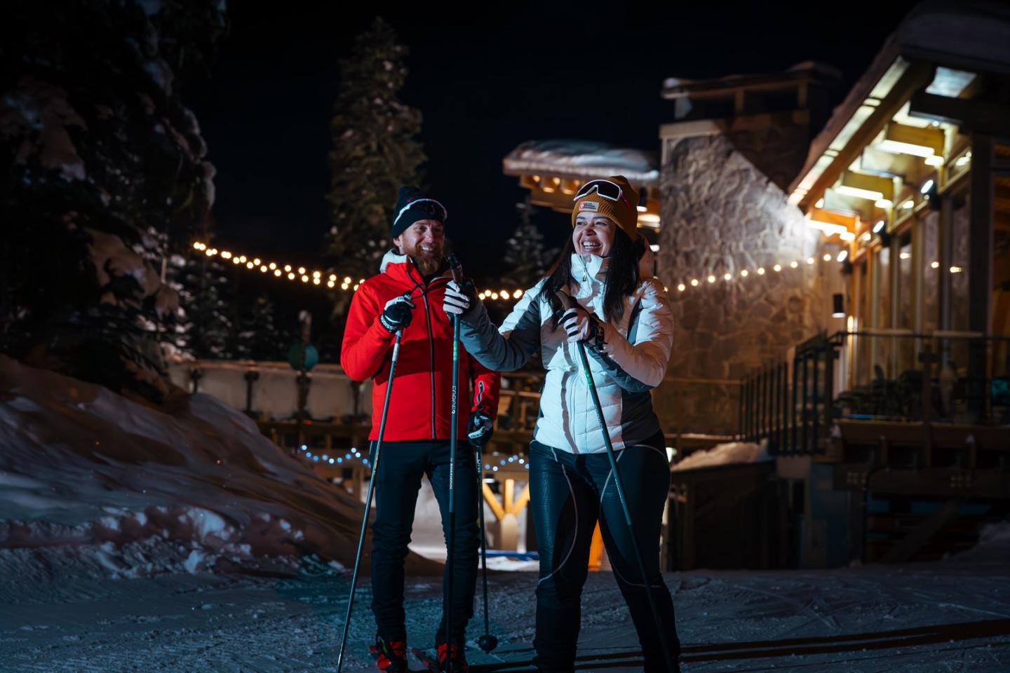 Two people in winter attire night skiing near a lit cabin.