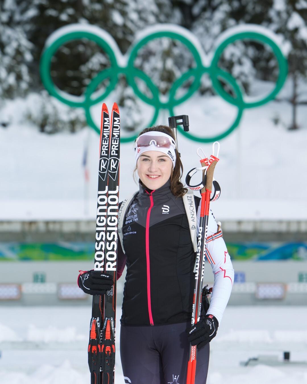 Athlete smiles with skis in snowy setting, Olympic rings in background.