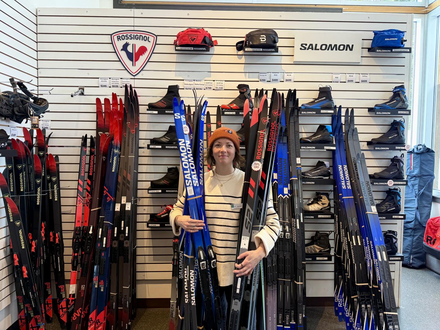 Person in a ski shop holding blue skis, surrounded by ski equipment.