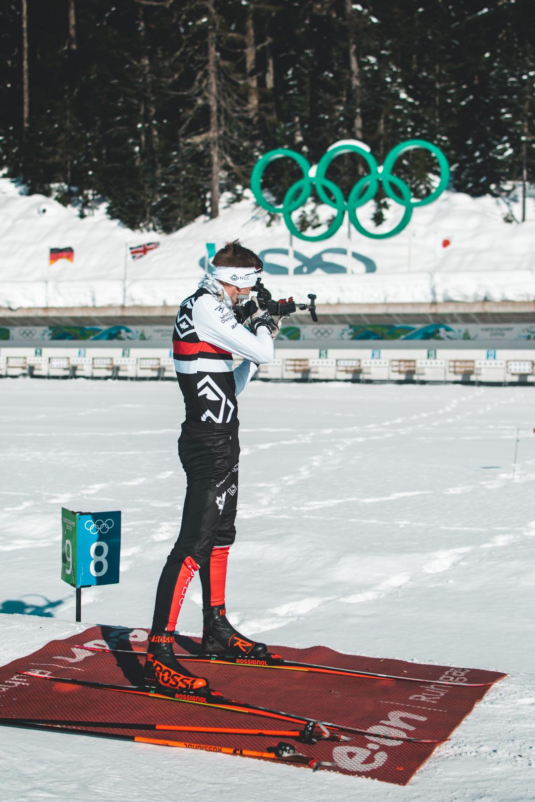 Biathlete aiming with a rifle on a snowy range, Olympic rings in the background.