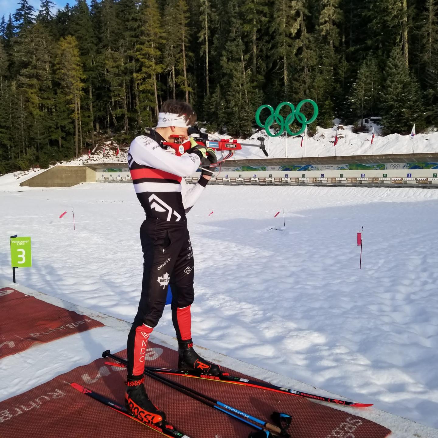 Cross-country skier aiming rifle at biathlon event in snowy forest setting.