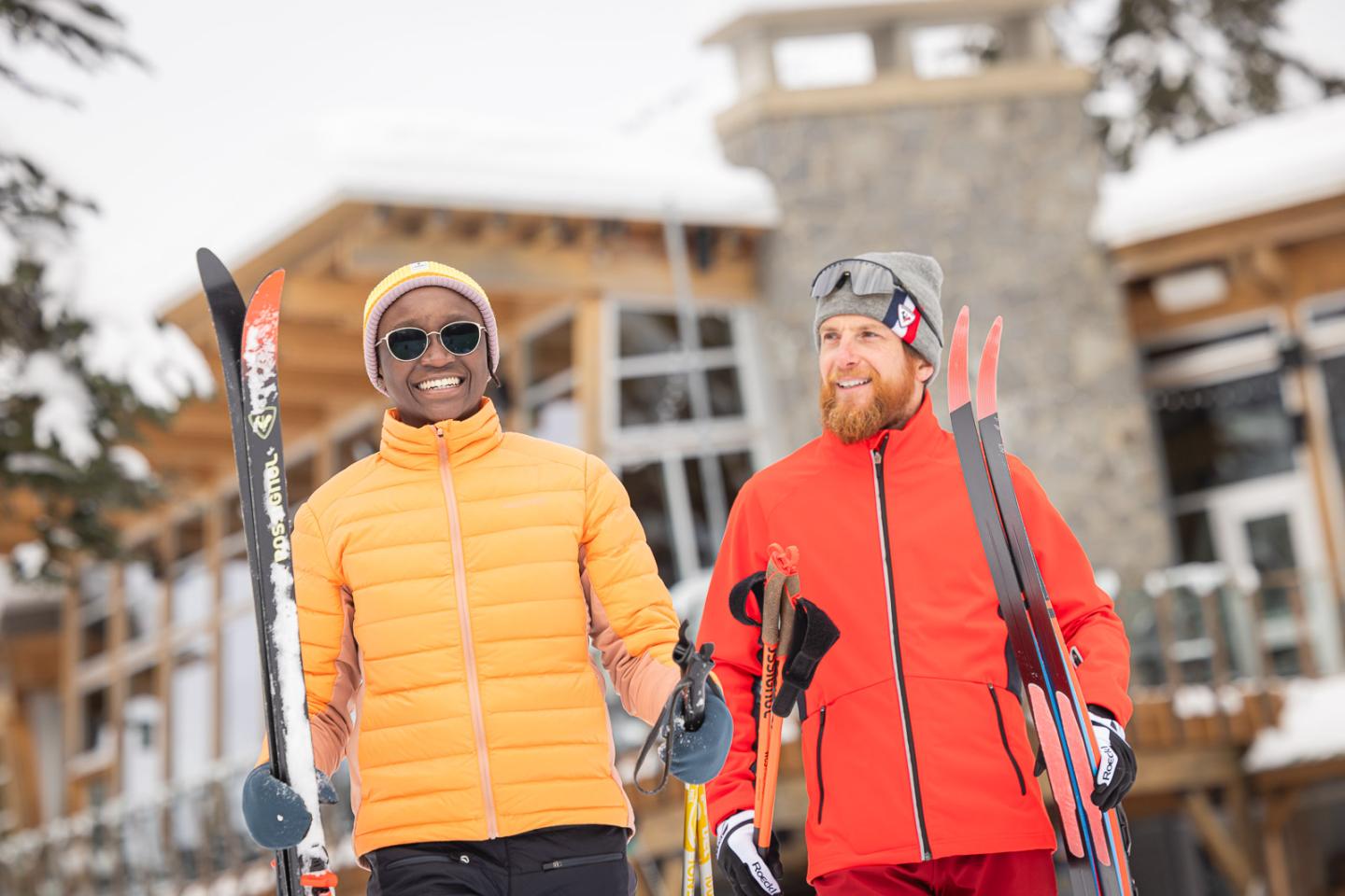 Two people in winter gear with skis, smiling outside a lodge.