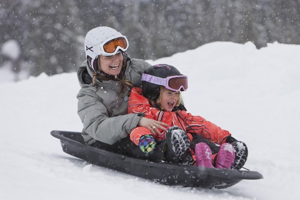 Sledding in snow, smiling woman and child in winter gear and goggles.