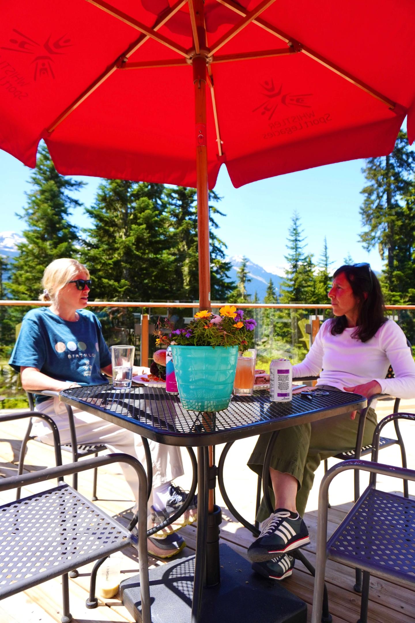 Two people sitting at a patio table under a red umbrella, surrounded by trees.