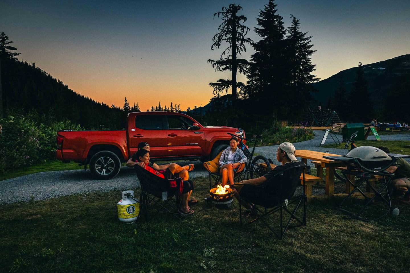 Campers by a fire near a red truck at sunset in the mountains.