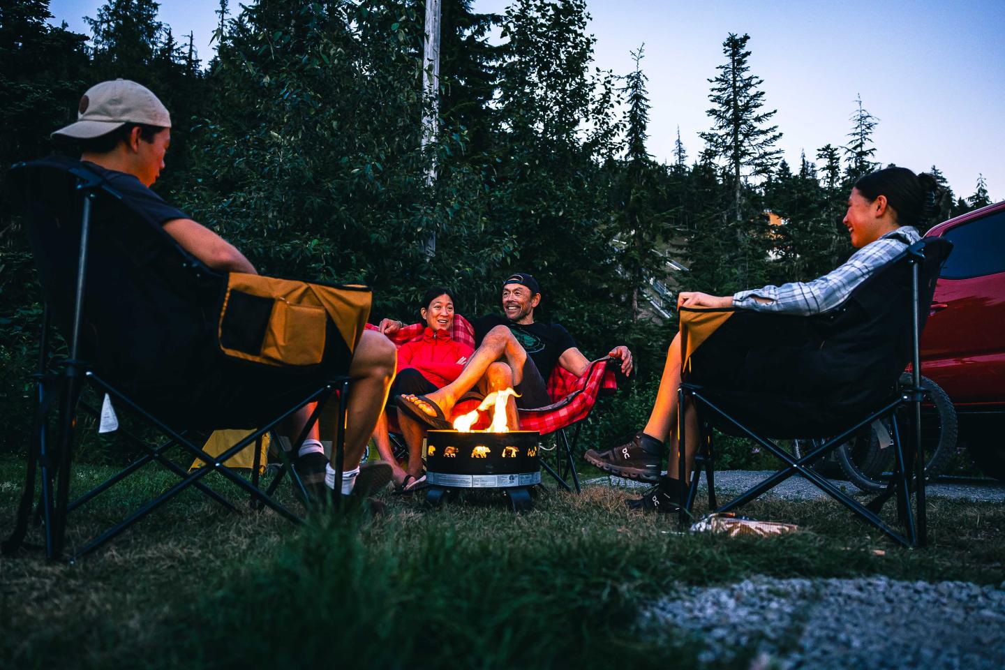 Four people sitting around a campfire at dusk in a forest.