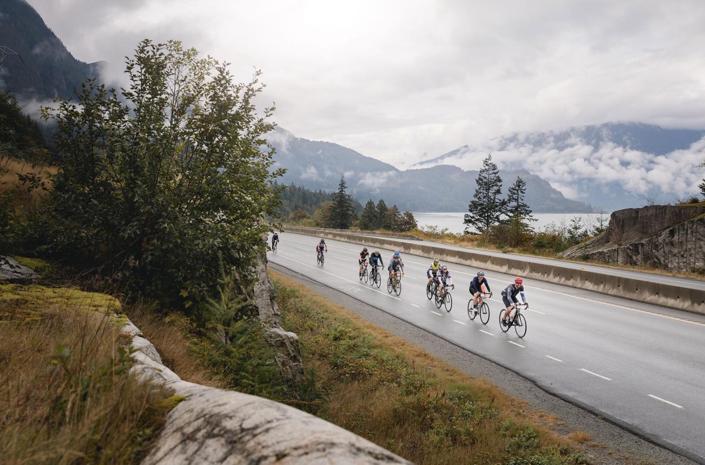 Cyclists ride along a wet mountain road beneath cloudy skies.